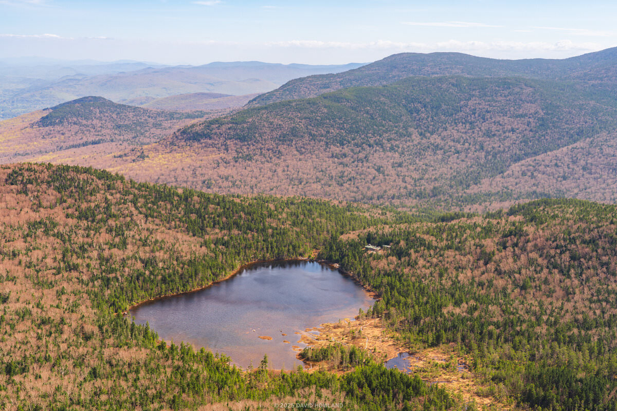 This landscape photograph captures a pristine mountain lake nestled within New England's rolling hills during spring, with the forested landscape displaying a mix of emerging green foliage and bare deciduous trees creating a patchwork of colors across the undulating terrain. The circular lake sits like a dark mirror in the valley below, surrounded by multiple ridgelines that fade into distant blue-gray mountain silhouettes under soft, diffused lighting.