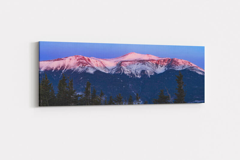 A panoramic morning alpenglow photograph of snow-covered Mount Washington and Tuckerman Ravine glowing in shades of pink and red against a deep blue pre-dawn sky, with the broad glacial bowl of Tuckerman Ravine clearly visible on the mountain's face, dark evergreen trees silhouetted in the foreground, and a small structure visible on the distant summit.
