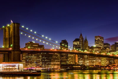 A panorama photo of a crescent moon rising over the Brooklyn Bridge and illuminated skyscrapers of downtown New Yok City at night.