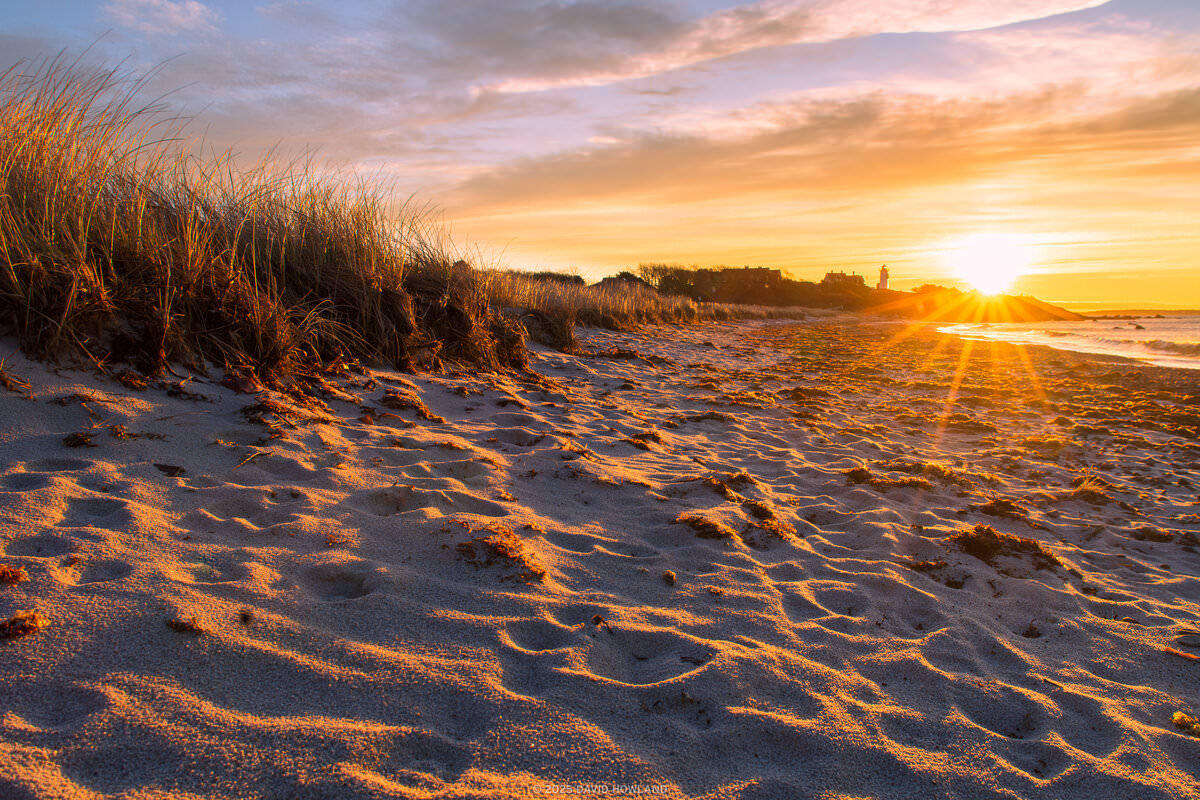 A photo of the sun rising behind Nobska Lighthouse and the sand and beach grass of Nobska Beach in Woods Hole, Massachusetts on Cape Cod.