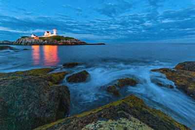 A lighthouse with red and white buildings sits on a rocky island at twilight, its warm light reflecting on calm blue water. Moss-covered rocks in the foreground frame the serene coastal scene under a cloudy evening sky.