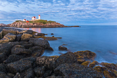 A photo of seaweed covered rocks on the coast of Maine with Nubble Lighthouse in the background.