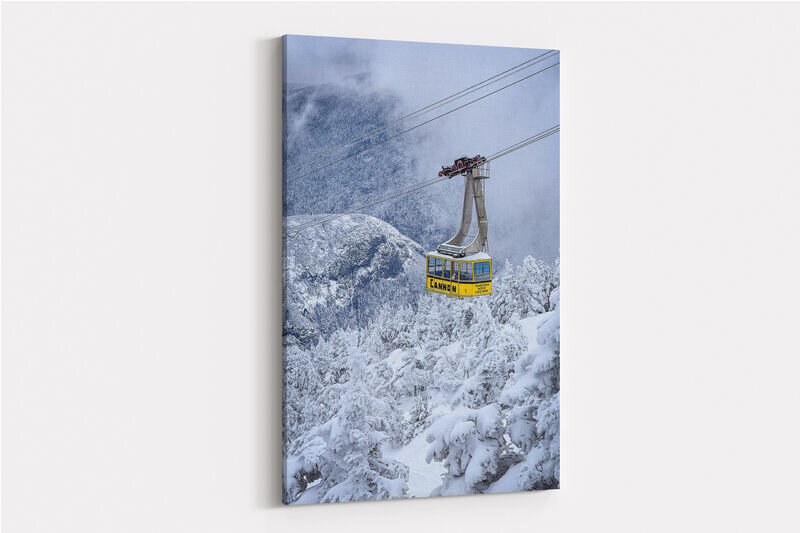 A bright yellow aerial tramway car ascends over a snow-covered mountain landscape with heavily frosted evergreen trees and misty peaks in the background at Cannon Mountain.