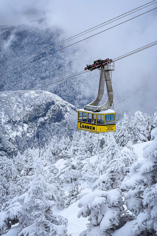 A bright yellow aerial tramway car ascends over a snow-covered mountain landscape with heavily frosted evergreen trees and misty peaks in the background at Cannon Mountain.