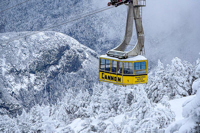 A bright yellow aerial tramway car ascends over a snow-covered mountain landscape with heavily frosted evergreen trees and misty peaks in the background at Cannon Mountain.