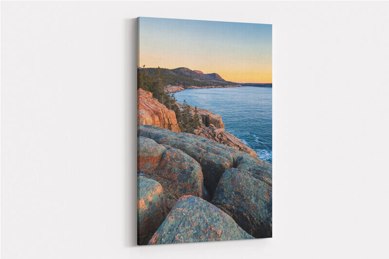 A sunrise view from rocky coastal cliffs showing lichen-covered granite boulders in the foreground, calm blue ocean waters, a tree-lined rocky shoreline, and distant forested mountains under a gradient sky transitioning from blue to golden yellow.