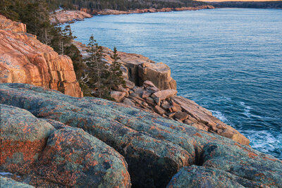 A sunrise view from rocky coastal cliffs showing lichen-covered granite boulders in the foreground, calm blue ocean waters, a tree-lined rocky shoreline, and distant forested mountains under a gradient sky transitioning from blue to golden yellow.