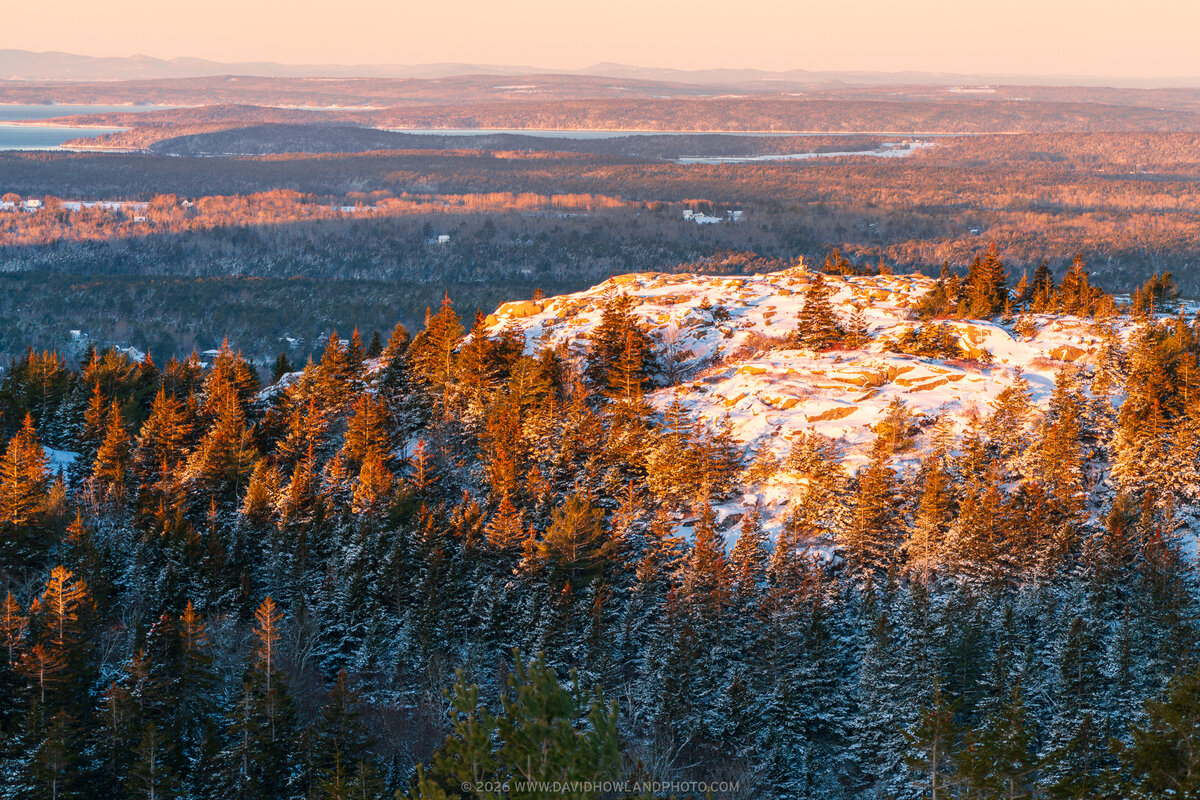 The snow-dusted rocky summit of Parkman Mountain in Acadia National Park glows golden in warm sunrise light, surrounded by evergreen trees and overlooking a vast winter landscape of forested hills, frozen lakes, and distant mountains under a soft pink sky.