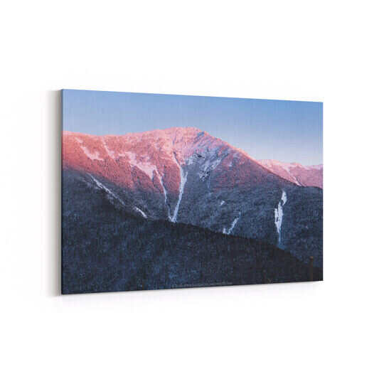 A winter sunset casts pink and purple light across the snow-covered peak of Mount Lincoln in New Hampshire's White Mountains, with evergreen forests covering the shadowed lower slopes and white avalanche chutes cutting through the mountainside.
