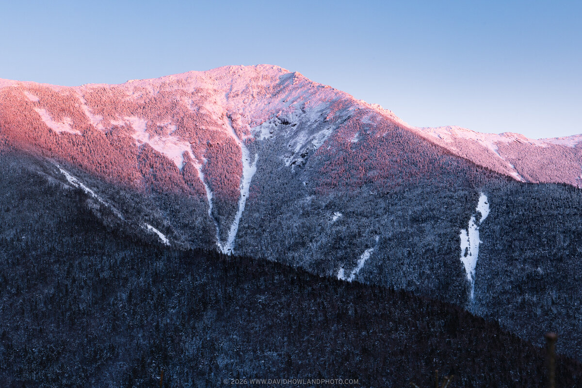 A winter sunset casts pink and purple light across the snow-covered peak of Mount Lincoln in New Hampshire's White Mountains, with evergreen forests covering the shadowed lower slopes and white avalanche chutes cutting through the mountainside.