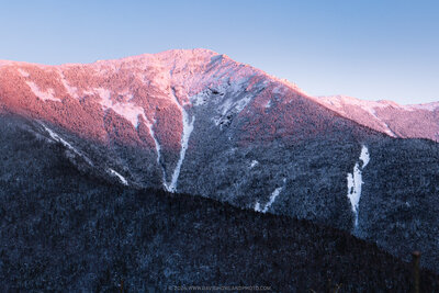 A winter sunset casts pink and purple light across the snow-covered peak of Mount Lincoln in New Hampshire's White Mountains, with evergreen forests covering the shadowed lower slopes and white avalanche chutes cutting through the mountainside.