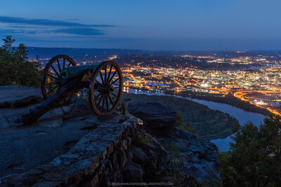 A Civil War-era cannon positioned on rocky fortifications at Point Park overlooks the illuminated city of Chattanooga at dusk, with the Tennessee River winding through the valley below and mountain silhouettes visible against the blue twilight sky.