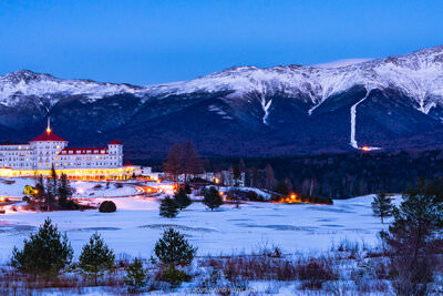 A panorama photo of the glowing orange lights of the Mount Washington Hotel building in front of the snow covered Presidential mountain range in New Hampshire.