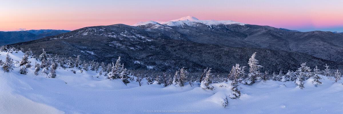 A panoramic winter sunset view from Mount Jackson in New Hampshire's Presidential Range, showing deep snow drifts and snow-laden evergreen trees in the foreground, with Mounts Eisenhower, Pierce, and Monroe visible across dark forested ridges, and snow-capped Mount Washington rising prominently against a glowing pink and lavender twilight sky.