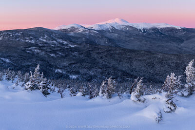A panoramic winter sunset view from Mount Jackson in New Hampshire's Presidential Range, showing deep snow drifts and snow-laden evergreen trees in the foreground, with Mounts Eisenhower, Pierce, and Monroe visible across dark forested ridges, and snow-capped Mount Washington rising prominently against a glowing pink and lavender twilight sky.