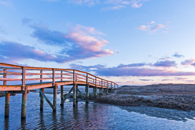 A photo of sunset lighting up the clouds in the sky over a boardwalk crossing a tidal channel at Ridgevale Beach in Chatham, MA.