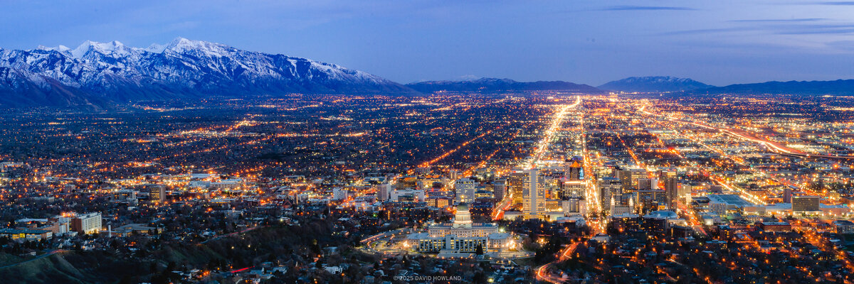 An elevated view from Ensign Peak of Salt Lake Valley at dusk, showing the illuminated city grid spreading across the valley floor with thousands of orange and white streetlights and buildings, backed by snow-covered mountains under a deep blue twilight sky.