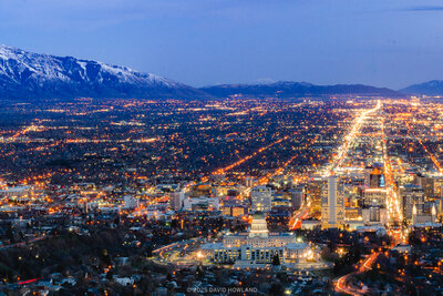 An elevated view from Ensign Peak of Salt Lake Valley at dusk, showing the illuminated city grid spreading across the valley floor with thousands of orange and white streetlights and buildings, backed by snow-covered mountains under a deep blue twilight sky.