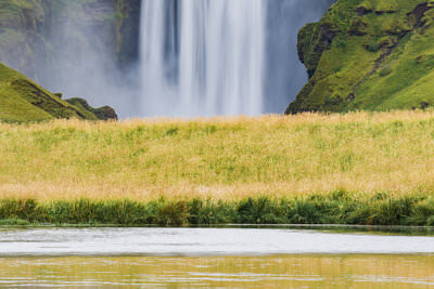 A photo of a towering waterfall reflected in the water surface in the green colored hills of Iceland.