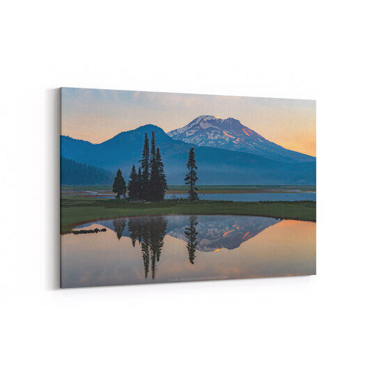 A smoky sunrise landscape at Sparks Lake, Oregon, featuring snow-capped South Sister glowing with warm orange light on its summit, framed by dark silhouetted evergreen trees on a grassy bank, with the entire scene — mountain, trees, and pastel sky — perfectly mirrored in the still foreground lake water.