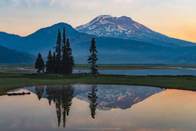 A smoky sunrise landscape at Sparks Lake, Oregon, featuring snow-capped South Sister glowing with warm orange light on its summit, framed by dark silhouetted evergreen trees on a grassy bank, with the entire scene — mountain, trees, and pastel sky — perfectly mirrored in the still foreground lake water.