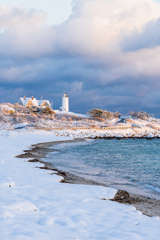 A snow-covered Nobska Lighthouse sits on a coastal bluff above blue ocean water and a snowy beach, bathed in warm golden sunset light against dramatic blue-gray storm clouds.