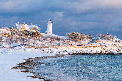 A snow-covered Nobska Lighthouse sits on a coastal bluff above blue ocean water and a snowy beach, bathed in warm golden sunset light against dramatic blue-gray storm clouds.