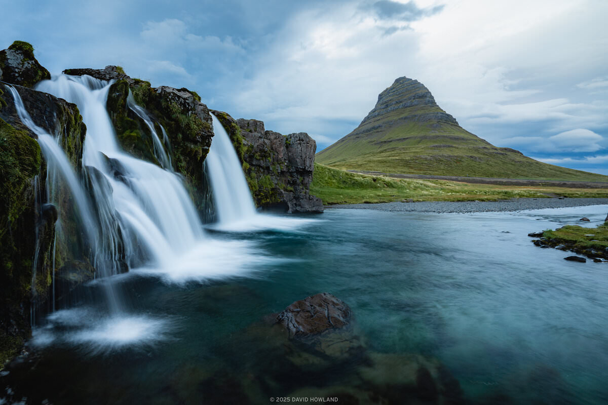 A landscape photograph showing Kirkjufellsfoss waterfall in the foreground with multiple white water cascades flowing over dark, moss-covered rocks into a turquoise pool. The distinctive pyramid-shaped Kirkjufell mountain rises prominently in the background against a cloudy blue sky. The scene includes green tundra vegetation in the middle ground and calm water extending toward the mountain base. The waterfall appears smooth and silky due to long exposure photography technique, while the overall composition captures the dramatic contrast between the flowing water and the static mountain landscape under an overcast sky.