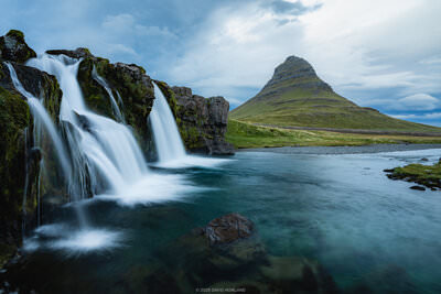 A landscape photograph showing Kirkjufellsfoss waterfall in the foreground with multiple white water cascades flowing over dark, moss-covered rocks into a turquoise pool. The distinctive pyramid-shaped Kirkjufell mountain rises prominently in the background against a cloudy blue sky. The scene includes green tundra vegetation in the middle ground and calm water extending toward the mountain base. The waterfall appears smooth and silky due to long exposure photography technique, while the overall composition captures the dramatic contrast between the flowing water and the static mountain landscape under an overcast sky.