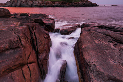 A sunset photo of waves crashing on the rocks of the coast of Maine with Nubble Lighthouse in the background.