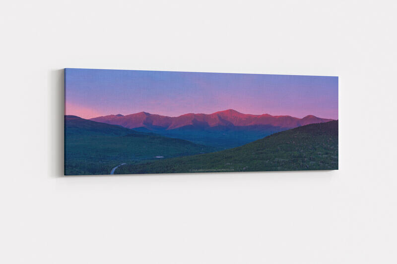 The Presidential Range glows with warm pink and red alpenglow at sunset, with the rocky peaks rising above dark forested valleys and green foothills under a purple and pink twilight sky.
