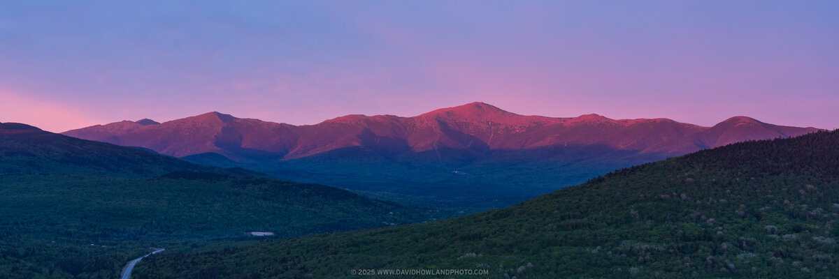 The Presidential Range glows with warm pink and red alpenglow at sunset, with the rocky peaks rising above dark forested valleys and green foothills under a purple and pink twilight sky.