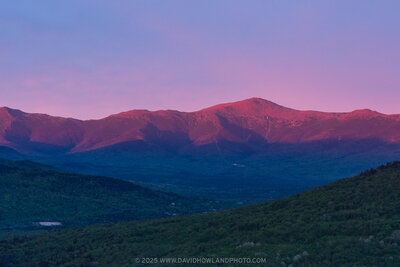 The Presidential Range glows with warm pink and red alpenglow at sunset, with the rocky peaks rising above dark forested valleys and green foothills under a purple and pink twilight sky.