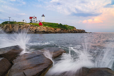 A sunset photo of waves crashing on the rocks of the coast of Maine with Nubble Lighthouse in the background.