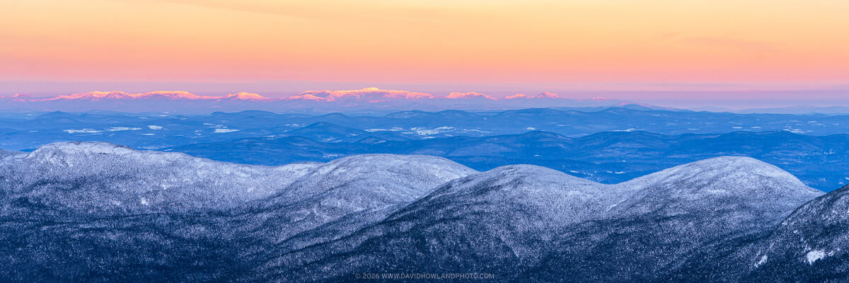 A panoramic winter sunrise landscape showing the snow-dusted Cannonball peaks in the foreground, rolling blue-toned hills in the middle distance, and Mount Mansfield and the Green Mountains glowing with warm alpenglow light on the far horizon beneath a vivid orange and pink sunrise sky.