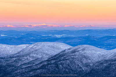 A panoramic winter sunrise landscape showing the snow-dusted Cannonball peaks in the foreground, rolling blue-toned hills in the middle distance, and Mount Mansfield and the Green Mountains glowing with warm alpenglow light on the far horizon beneath a vivid orange and pink sunrise sky.
