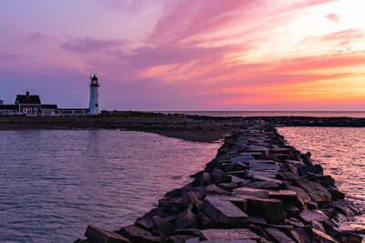 A photo of a rock jetty and Scituate Lighthouse in front of a colorful sunrise.