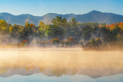 A pond at sunrise with dense golden mist hovering over the water's surface and partially obscuring the shoreline. Beyond the mist, a forest displays autumn foliage in orange, yellow, and green, with blue mountains visible in the background.