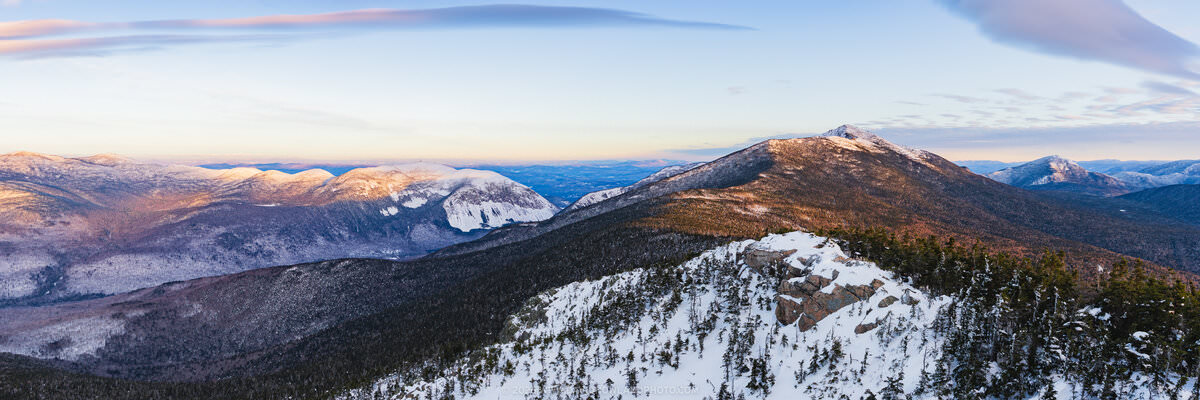 A panoramic winter sunrise photograph taken from a snow-covered rocky summit, looking across Franconia Notch with snow-dusted evergreen trees in the foreground, rocky outcroppings at center, and the illuminated peaks of Cannon Mountain, Little Haystack Mountain, and Mount Lincoln bathed in warm golden-pink sunrise light against a pale blue sky with wispy clouds.