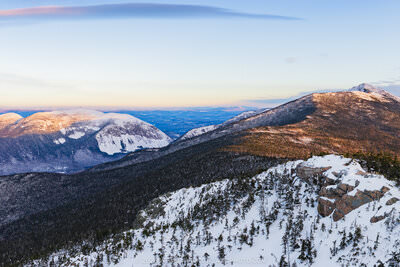 A panoramic winter sunrise photograph taken from a snow-covered rocky summit, looking across Franconia Notch with snow-dusted evergreen trees in the foreground, rocky outcroppings at center, and the illuminated peaks of Cannon Mountain, Little Haystack Mountain, and Mount Lincoln bathed in warm golden-pink sunrise light against a pale blue sky with wispy clouds.