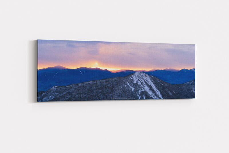 A panoramic winter sunrise photograph showing snow-dusted Mount Flume in the foreground and the Sandwich Range silhouetted in deep blue against a glowing pink, lavender, and orange sky with the sun just cresting the distant ridgeline.