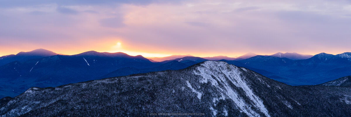 A panoramic winter sunrise photograph showing snow-dusted Mount Flume in the foreground and the Sandwich Range silhouetted in deep blue against a glowing pink, lavender, and orange sky with the sun just cresting the distant ridgeline.