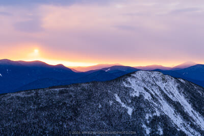 A panoramic winter sunrise photograph showing snow-dusted Mount Flume in the foreground and the Sandwich Range silhouetted in deep blue against a glowing pink, lavender, and orange sky with the sun just cresting the distant ridgeline.
