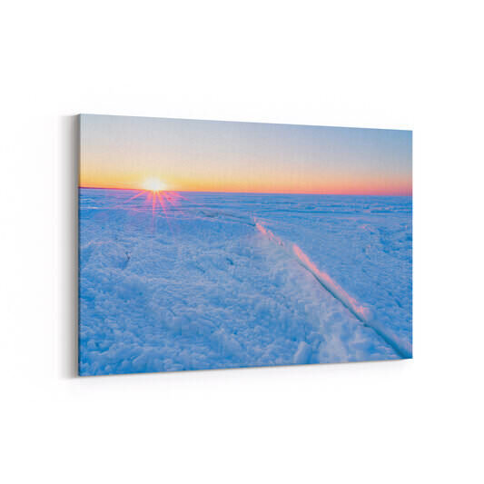 A winter sunset casts golden-pink light across a frozen beach covered in chunky, textured ice formations, with the sun low on the horizon creating dramatic rays of light over the icy landscape.
