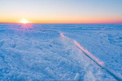 A winter sunset casts golden-pink light across a frozen beach covered in chunky, textured ice formations, with the sun low on the horizon creating dramatic rays of light over the icy landscape.