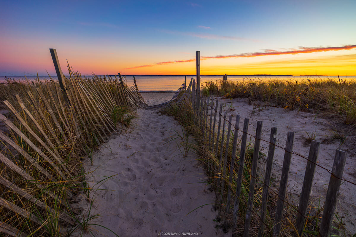 A photo of a battered wooden fence marking a path through the dunes of Surf Drive Beach at sunset in Falmouth, Massachusetts.