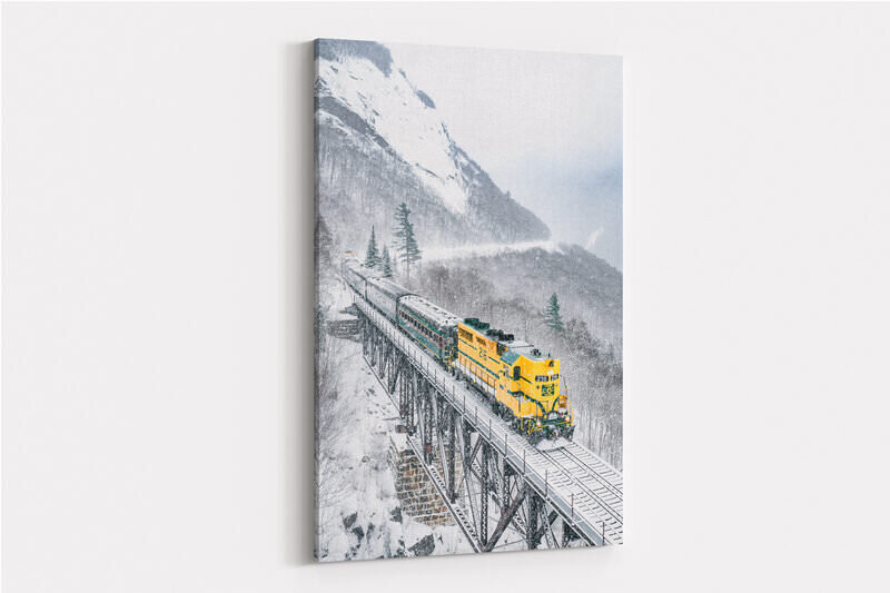 A bright yellow locomotive pulling green passenger cars crosses a tall steel trestle bridge through a snowy mountain valley during heavy snowfall, with steep, snow-covered slopes and evergreen trees visible in the background.