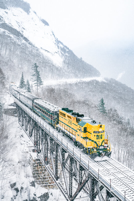 A bright yellow locomotive pulling green passenger cars crosses a tall steel trestle bridge through a snowy mountain valley during heavy snowfall, with steep, snow-covered slopes and evergreen trees visible in the background.
