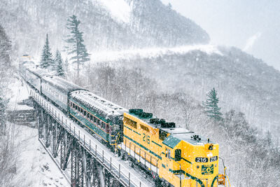 A bright yellow locomotive pulling green passenger cars crosses a tall steel trestle bridge through a snowy mountain valley during heavy snowfall, with steep, snow-covered slopes and evergreen trees visible in the background.