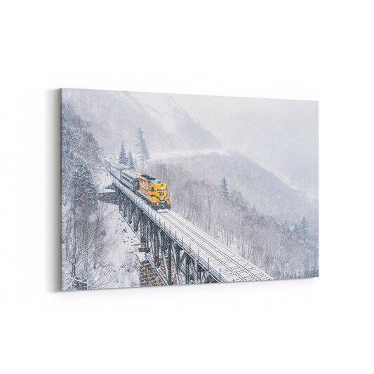 A bright yellow locomotive pulls passenger cars across a snow-covered railroad trestle bridge through a misty winter mountain landscape at Willey Brook Bridge in Crawford Notch State Park, with frost-laden trees and steep mountainsides visible through falling snow.